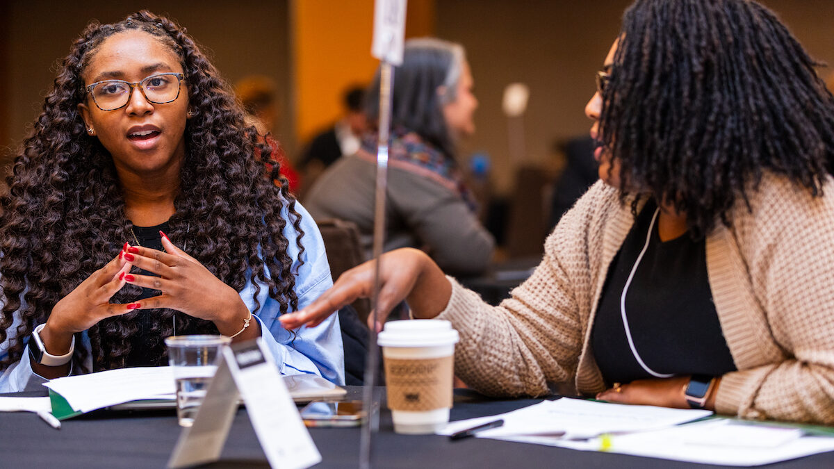 Two women sit at a table in discussion with papers and coffee cups as they map out strategic visioning. Both are engaged in conversation.